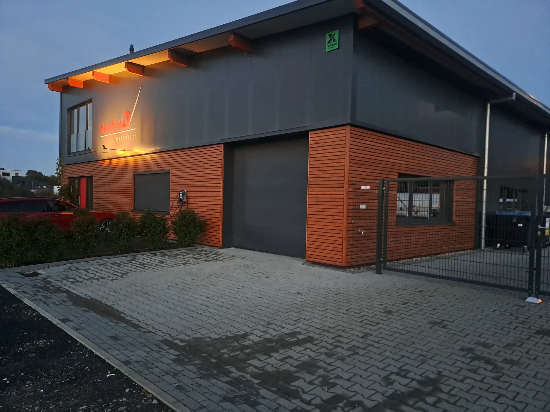 The Maaßen joinery in Mönchengladbach at dusk: timber façade and dark metal cladding, with illuminated lettering above the hall door.