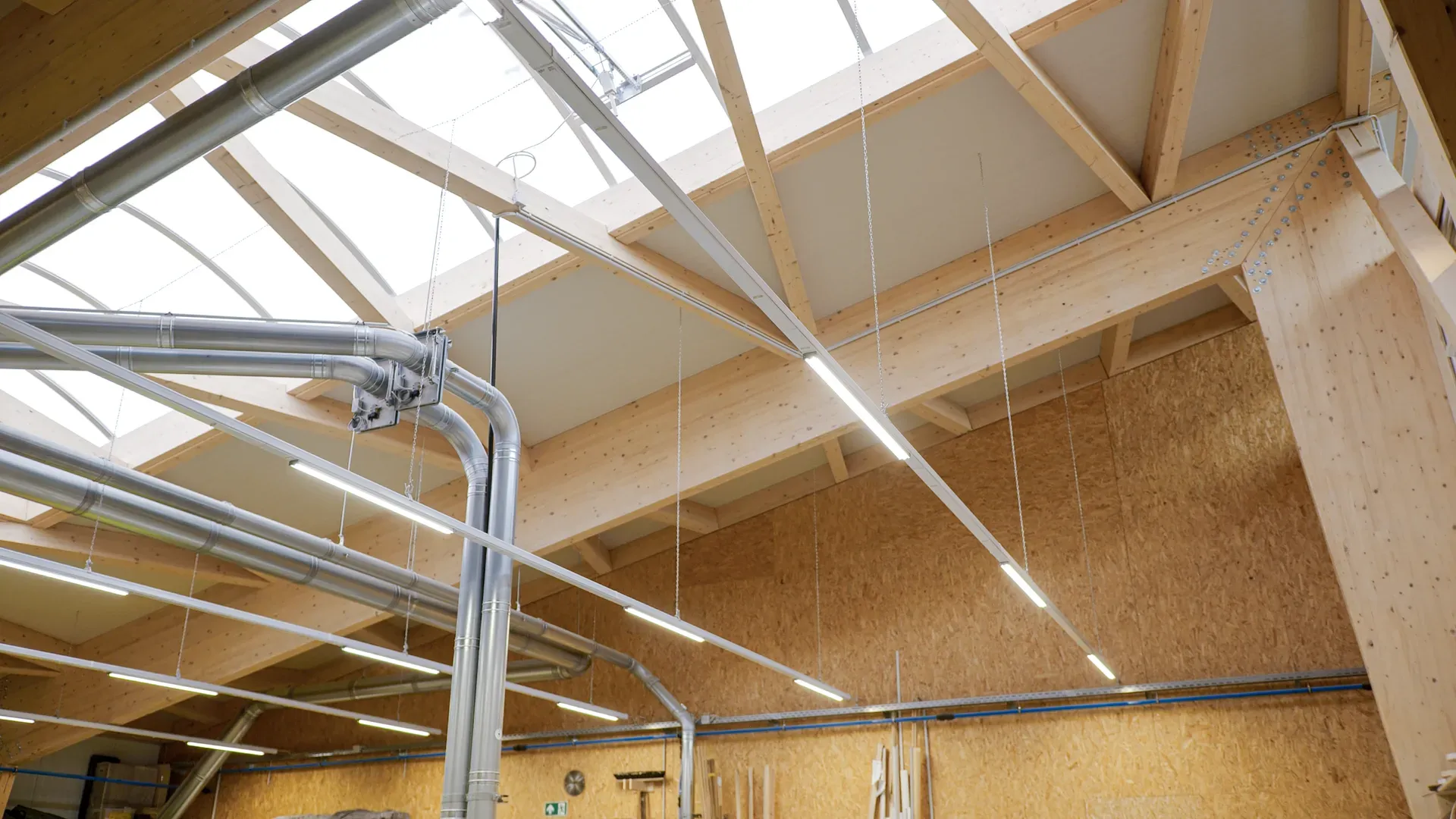 Roof structure of the 2Tischler hall in Aachen with a ridge light strip, exposed timber trusses, an extraction system and OSB walls.