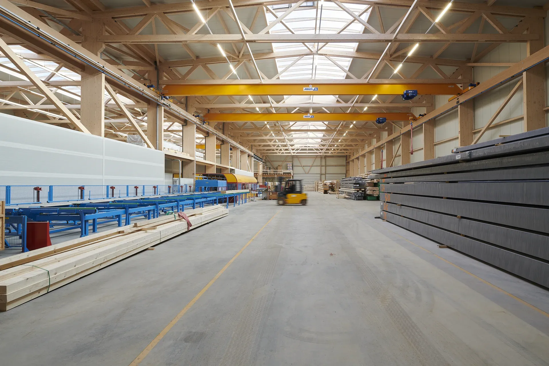 Central aisle of the X-CAM production hall in Kall with timber lattice trusses, two yellow crane runways and a ridge light.