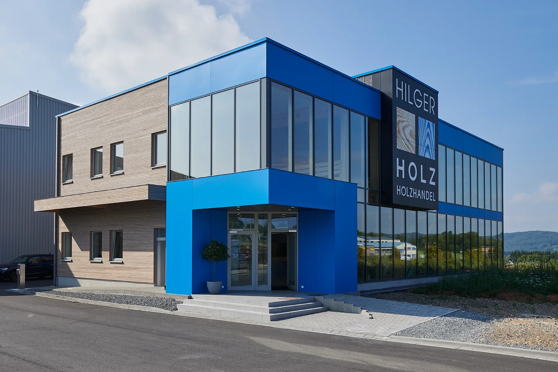 Exterior view of the Hilger Holz showroom in Kall, with a blue façade, timber cladding, a glazed building corner and the Hilger Holz Holzhandel lettering.